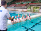 A swimming teacher is giving instructions. 7 children are sitting by the poolside in swimming costume with their feet in water. 2 children are in pool.