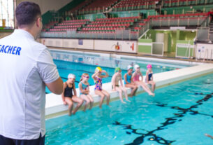 A swimming teacher is giving instructions. 7 children are sitting by the poolside in swimming costume with their feet in water. 2 children are in pool.